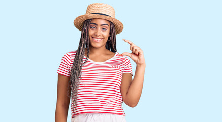 Obraz na płótnie Canvas Young african american woman with braids wearing summer hat smiling and confident gesturing with hand doing small size sign with fingers looking and the camera. measure concept.