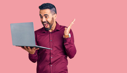 Young hispanic man holding laptop celebrating victory with happy smile and winner expression with raised hands