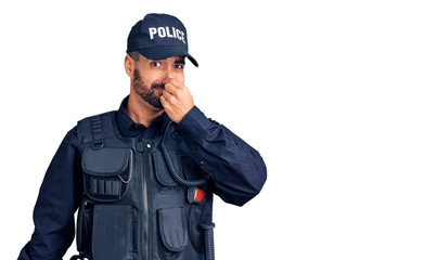Young hispanic man wearing police uniform smelling something stinky and disgusting, intolerable smell, holding breath with fingers on nose. bad smell