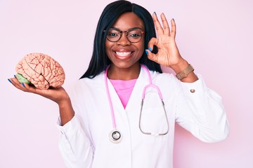 Young african american woman wearing doctor coat holding brain doing ok sign with fingers, smiling...