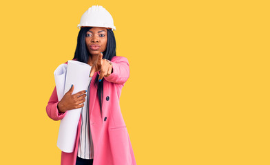 Young african american woman wearing architect hardhat holding blueprints pointing with finger to the camera and to you, confident gesture looking serious