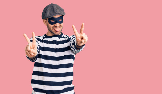 Young handsome man wearing burglar mask smiling with tongue out showing fingers of both hands doing victory sign. number two.