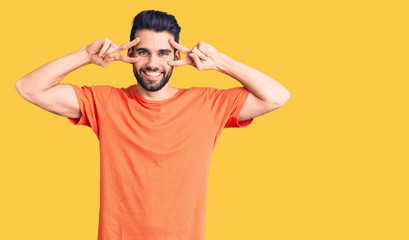 Young handsome man with beard wearing casual t-shirt doing peace symbol with fingers over face, smiling cheerful showing victory