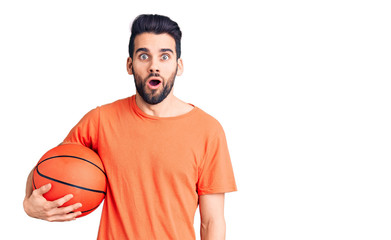 Fototapeta premium Young handsome man with beard hoilding basketball ball scared and amazed with open mouth for surprise, disbelief face