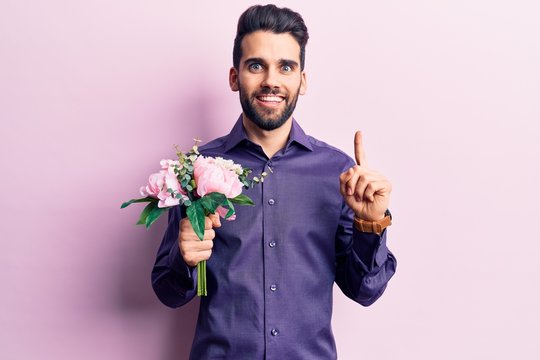 Young handsome man with beard holding bouquet of flowers smiling with an idea or question pointing finger with happy face, number one