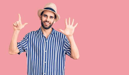 Young handsome man with beard wearing summer hat and striped shirt showing and pointing up with fingers number seven while smiling confident and happy.