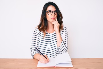 Young beautiful brunette woman writing book sitting on the table serious face thinking about question with hand on chin, thoughtful about confusing idea