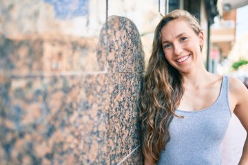 Young blonde woman smiling happy leaning on the wall at street of city