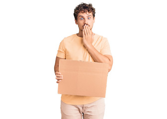 Young handsome man with curly hair holding blank cardboard empty banner covering mouth with hand,...