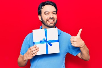 Young hispanic man holding gift smiling happy and positive, thumb up doing excellent and approval sign