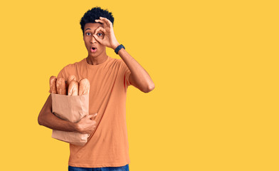 Young african american man holding paper bag with bread doing ok gesture shocked with surprised face, eye looking through fingers. unbelieving expression.