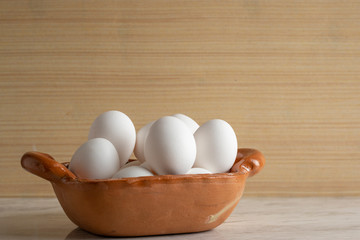 Vasija de barro llena de huevos blancos, sobre una barra de mármol y fondo de madera. Con espacio para publicidad en la parte superior.