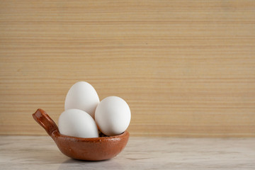 Cazuela de barro con huevos blancos, sobre una barra de mármol y fondo de madera. Con espacio para publicidad en la parte superior.