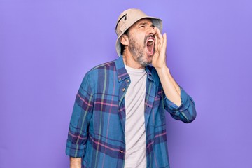 Middle age handsome man wearing casual shirt and hat over isolated purple background shouting and screaming loud to side with hand on mouth. Communication concept.