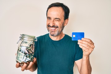 Middle age hispanic man holding credit car and jar with dollars winking looking at the camera with...
