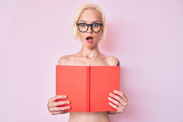 Young blonde woman with tattoo standing shirtless reading book afraid and shocked with surprise and amazed expression, fear and excited face.