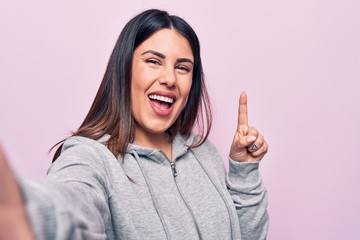 Young beautiful brunette sporty woman make selfie by the camera over pink background smiling with an idea or question pointing finger with happy face, number one