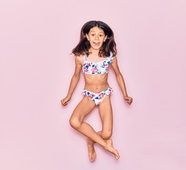 Adorable hispanic child girl on vacation wearing bikini surprised with open mouth. Jumping over isolated pink background - obrazy, fototapety, plakaty