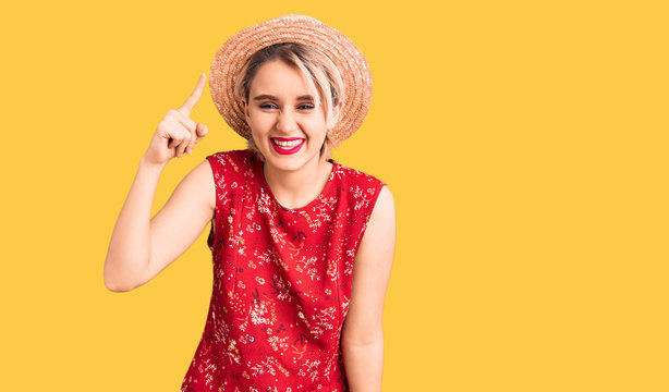 Young beautiful blonde woman wearing summer hat pointing finger up with successful idea. exited and happy. number one.