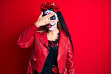 Woman wearing day of the dead costume over red peeking in shock covering face and eyes with hand, looking through fingers with embarrassed expression.