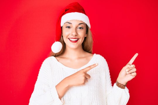 Young brunette woman wearing christmas hat smiling and looking at the camera pointing with two hands and fingers to the side.