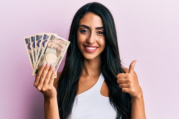 Beautiful hispanic woman holding japanese yen banknotes smiling happy and positive, thumb up doing excellent and approval sign