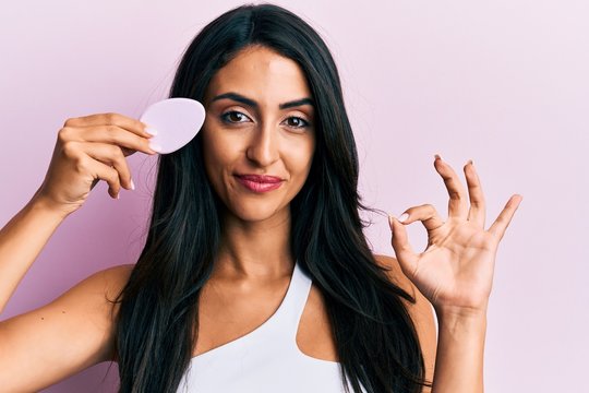 Beautiful Hispanic Woman Holding Makeup Sponge Doing Ok Sign With Fingers, Smiling Friendly Gesturing Excellent Symbol