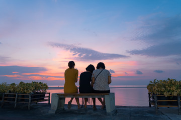 Fototapeta na wymiar Three asian girls sit on a bench by the sea and watch the sunset