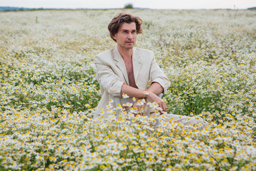 Tall handsome man sitting on a white chair in camomile flowers field