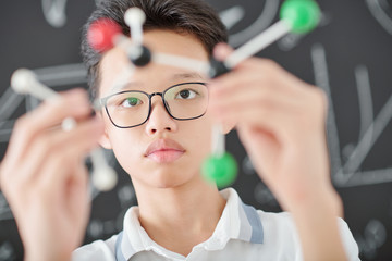 Intelligent serious Asian schoolboy in glasses examining plastic model of chemical element in his...