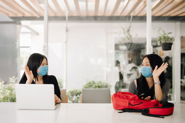 Asian two women greeting together wearing a mask in social distancing in office or classroom for prevention COVID-19 Virus. Social distancing concept.