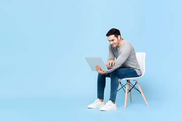 Portrait of smiling young handsome Caucasian man sitting while working on his laptop in isolated studio blue background