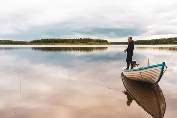 Foto op Canvas An elderly man is fishing from a boat near the shore on the lake © Sergey + Marina