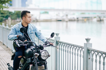 Handsome cool Asian man in denim jacket standing next to motorcycle and looking at river