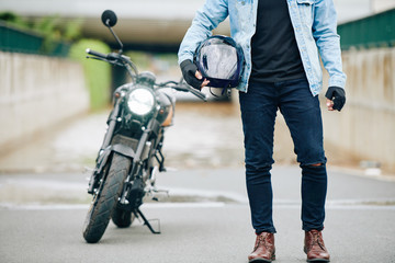 Naklejka premium Przycięty obraz motocyklisty w dżinsach i skórzanych butach, trzymając kask, jego motocykl jest w tle