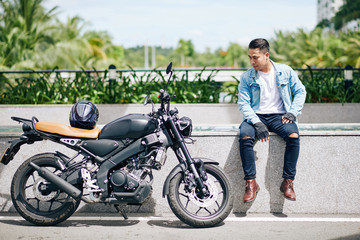 Fototapeta na wymiar Proud Vietnamese biker sitting on parapet and looking at his motorcycle