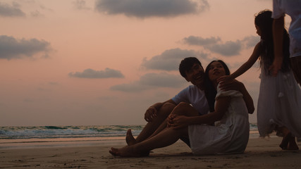Asian young happy family enjoy vacation on beach in evening. Dad, mom and kid sit, hug relax together near sea when looking silhouette sunset. Lifestyle travel holiday vacation summer concept.