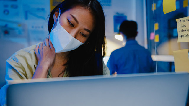 Stressed Tired Young Asia Woman Wear Face Mask Using Laptop Hard Work Having Office Syndrome , Neck Pain, While Working Overtime At Office. Working From Home Overload At Night, Social Distancing.