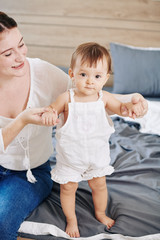 Smiling woman supporting her adorable little daughter learning how to walk on bed