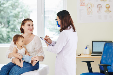 Obraz premium Smiling mother of adorable little girl answering questions of pediatrician in medical mask