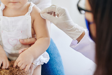 Doctor in rubber gloves wiping injection site on arm of little girl before vaccinate her for...