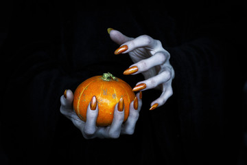 Small pumpkin in witches knotty hand. Scary undead pale fingers with long sharp claws in the dark,...