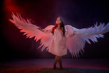 Small young model girl with white wings posing and dancing in dark black studo during photoshoot with flour or dust and colour light. Actress in performance about struggle between good and evil.
