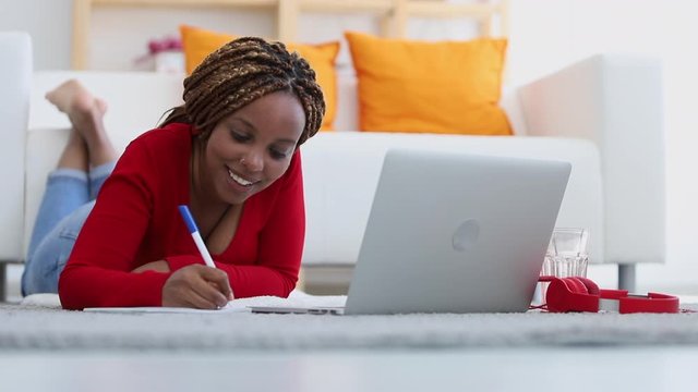 Beautiful young girl writing and using laptop while lying on floor at apartment room spbi. Front view of African American woman making notes in notebook and looking at computer screen with smile in