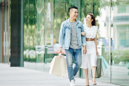 Happy pretty young Asian woman looking on her boyfriend when they are walking outdoors after shopping