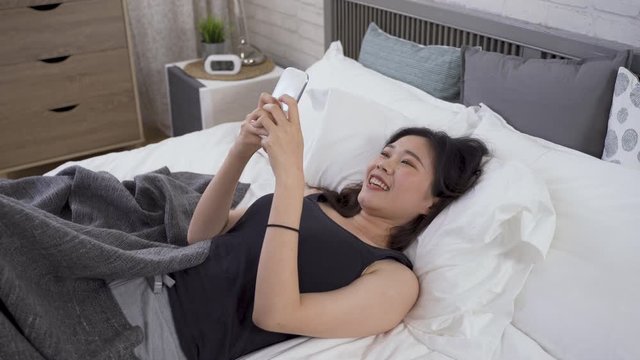 relaxing holiday morning asian woman holding phone is texting back to her boyfriend before getting out of bed. communication, technology and lifestyle