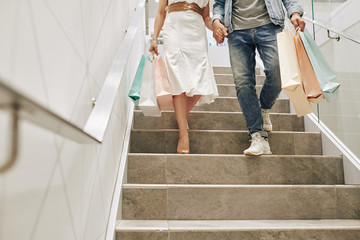 Cropped image of young couple with shopping bags holding hands when walking down the stairs after shopping in mall