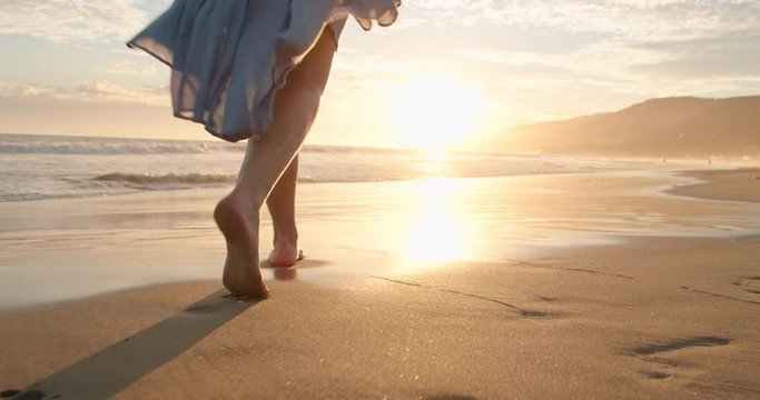 Slow motion woman feet walking barefoot by beach at golden sunset leaving footprints in sand. Female tourist on summer vacation in Malibu, California, USA. Woman in beautiful waving dress at sunset