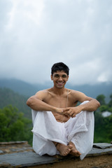 young shredded man sitting wearing a dhoti and glasses with blurred mountains in the background.