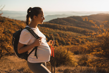 Pregnant woman nordic walking with trekking sticks on hill with beautiful autumn mountain view. Pregnancy activity and healthy lifestyle in maternity time. Travel concept.